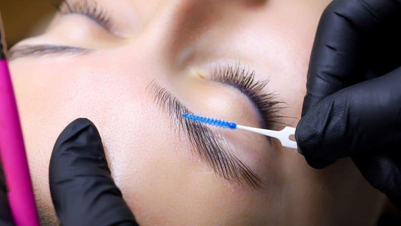 A closeup on a person's eyebrows while a gloved hand runs a spoolie through the hairs.