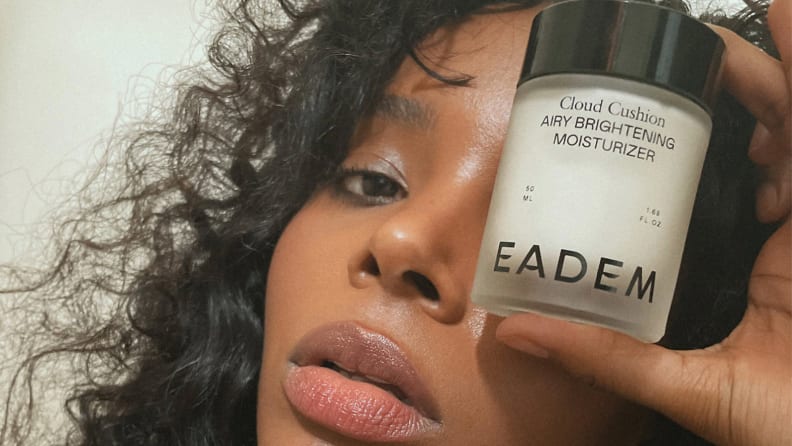 A person holding a jar of moisturizer up to their eye.